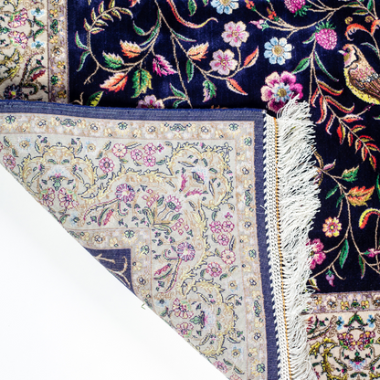 Navy silk rug with floral, bird, and vine motifs in pink, green, and multicolour tones, bordered in ivory.

