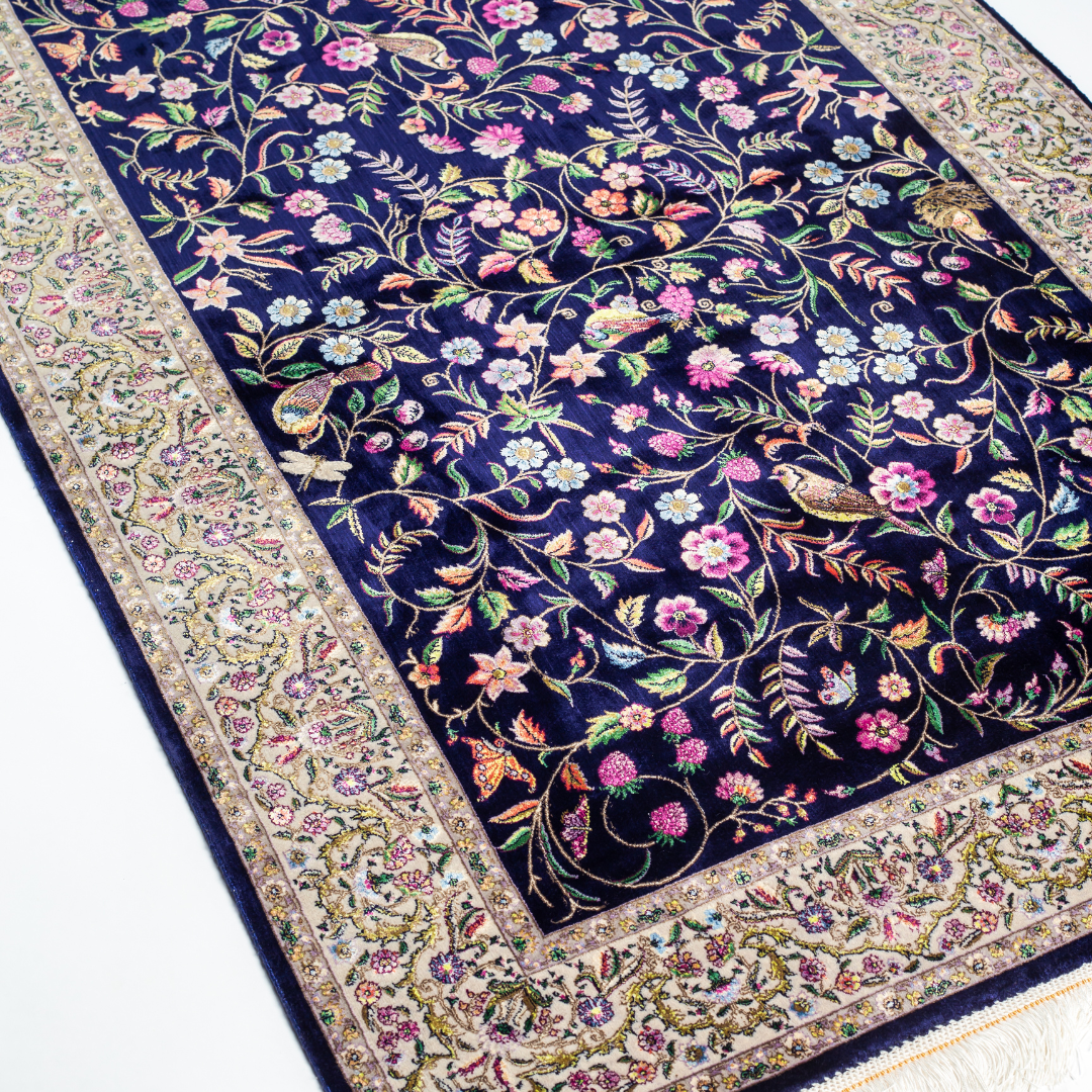 Navy silk rug with floral, bird, and vine motifs in pink, green, and multicolour tones, bordered in ivory.

