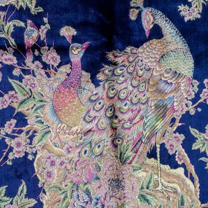 Luxurious silk rug with peacock pair and blooming florals on a navy base. Handwoven in soft hues with intricate botanical detailing.

