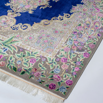 Luxurious silk rug with peacock pair and blooming florals on a navy base. Handwoven in soft hues with intricate botanical detailing.

