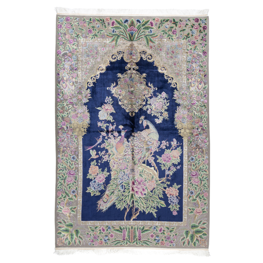 Luxurious silk rug with peacock pair and blooming florals on a navy base. Handwoven in soft hues with intricate botanical detailing.

